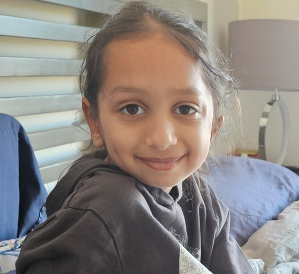 A photo SickKids patient Chitra at home, smiling and sitting on a bed