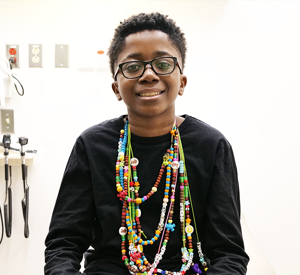 A portrait of Daniel with his arms crossed, looking strong and serious. He’s wearing colourful strands of bravery beads and a hospital gown.