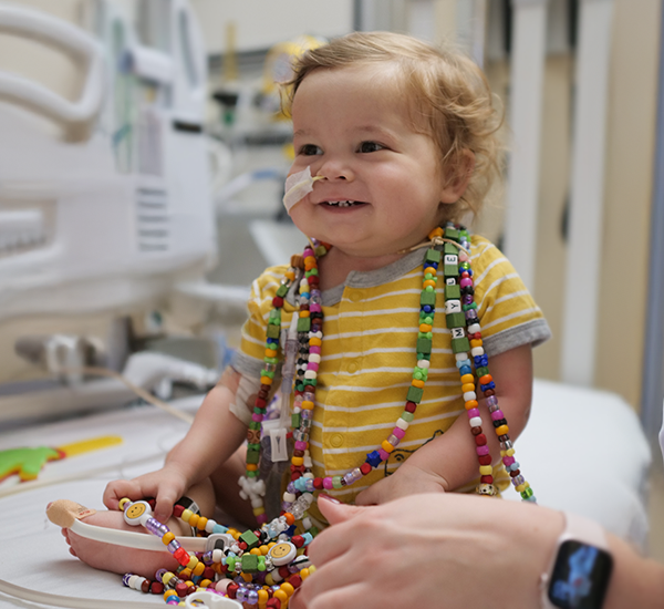 Myles at SickKids. He’s smiling and looking off camera, and wearing strands of colourful bravery beads. He has a feeding tube in his nose.