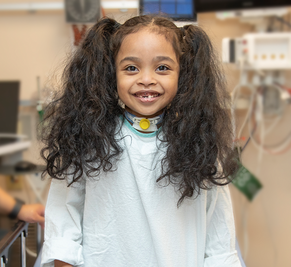 Noah standing beside a bed at SickKids. She’s smiling and wearing a hospital gown. 