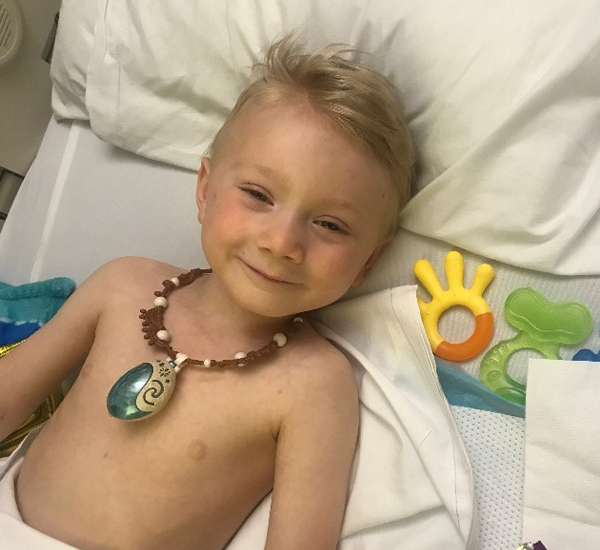 Austin at SickKids. He’s lying on a bed, wearing a necklace and smiling. 