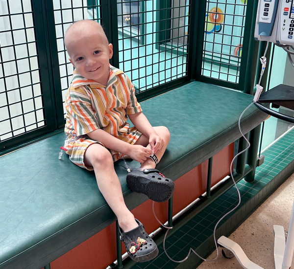 Sam sitting on a bench at SickKids, smiling. There is an IV pole next to him.