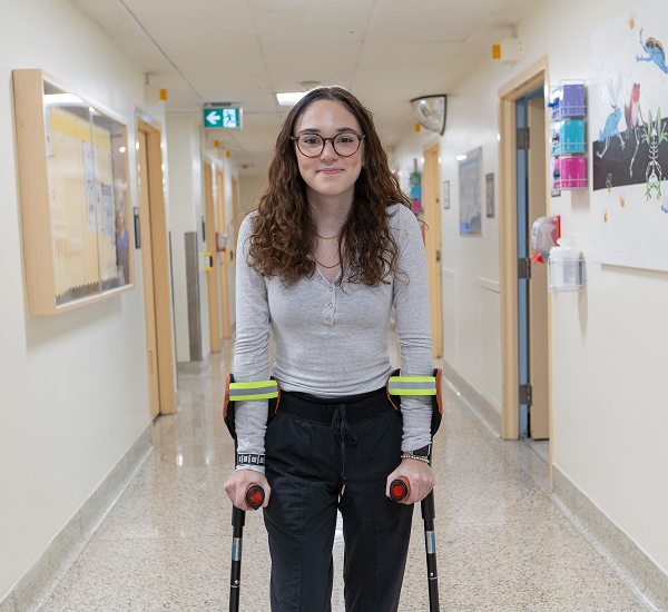 Hope smiling, standing in a hallway at SickKids with her crutches.