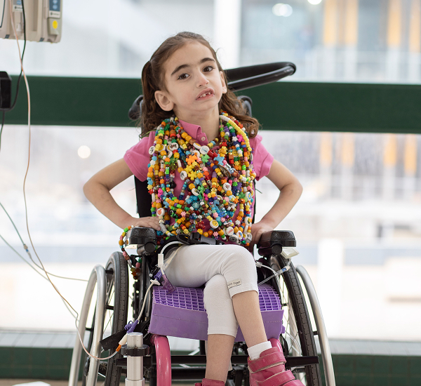 Adina at SickKids, sitting in her wheelchair and smiling, wearing colorful bravery beads necklaces.