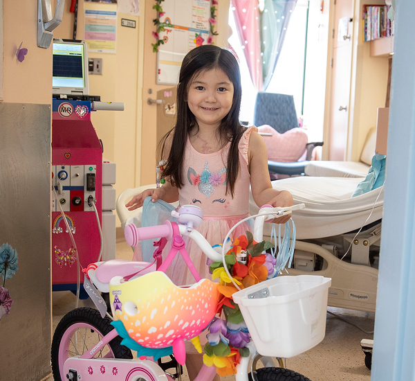 Harper at SickKids, smiling beside her decorated pink bike in her hospital room.