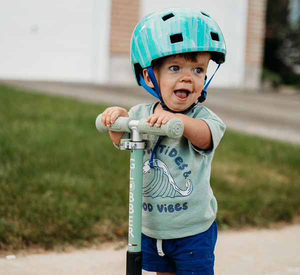 Lincoln outside, smiling while riding his scooter and wearing a helmet.