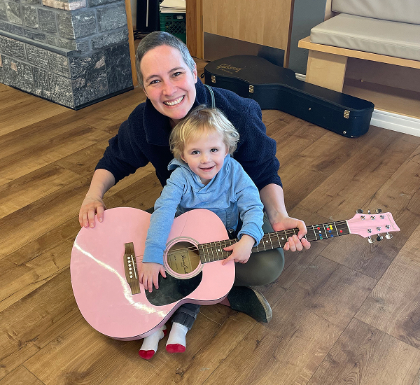 William at home, smiling while holding a pink guitar with a caregiver behind him.