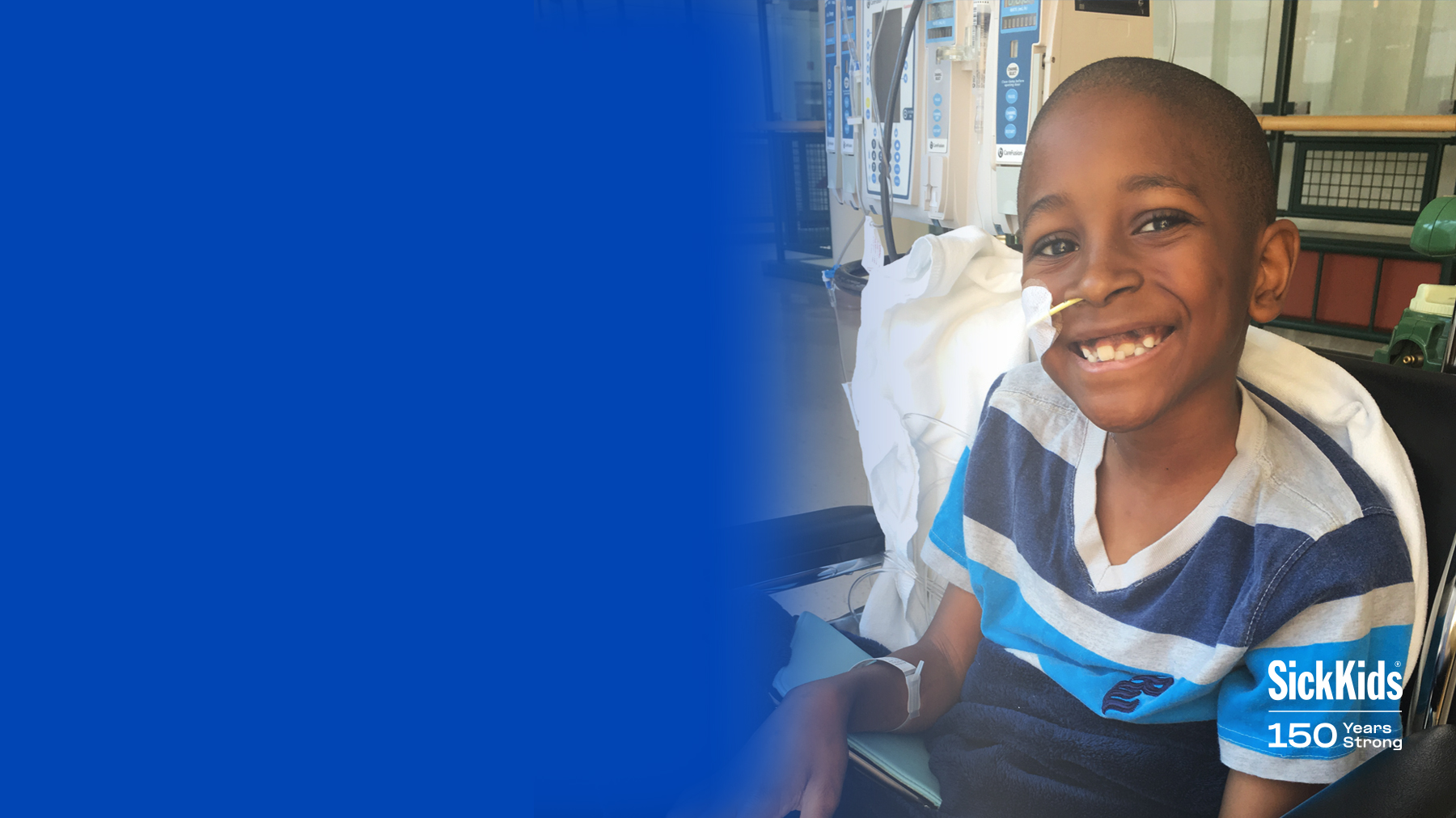 Tajaun at SickKids sitting in a chair. He’s smiling, wearing a blue and white striped T-shirt. He has a feeding tube in his nose.