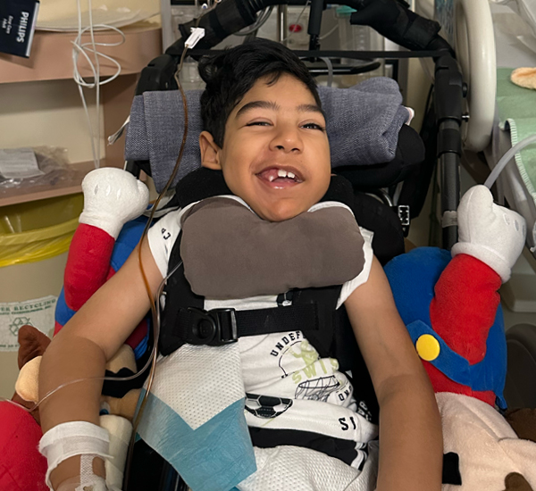 Shaan at SickKids. He’s smiling, and in his wheelchair surrounded by stuffed toys. There is an IV in his hand.