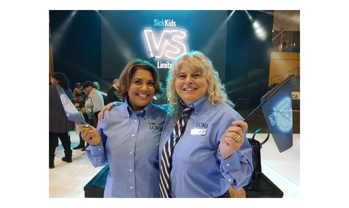 Two women wearing LCBO shirts and waving SickKids VS flags
