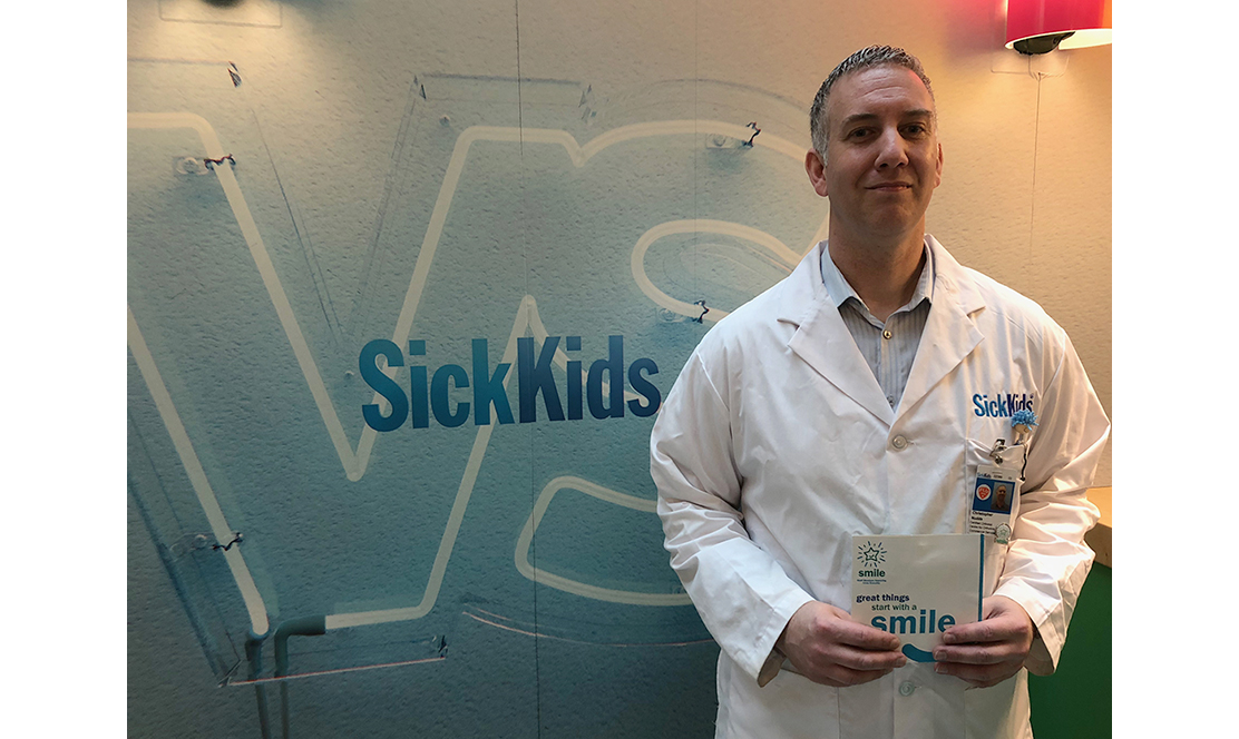 Man in lab coat standing next to SickKids VS logo
