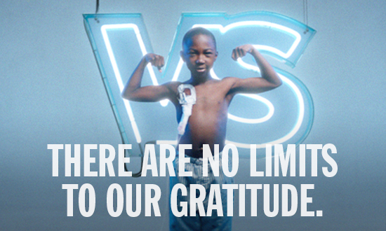 A young boy with dark skin and very short dark hair stands in front of a Sick Kids VS neon sign. The Sign is blue and white. The boy is flexing his biceps. On his chest is an affixed medical port. He is wearing blue and white striped pants. Text superimposed over the image reads: There are no limits to our gratitude.
