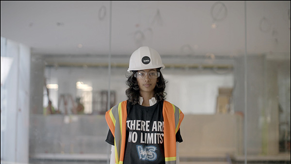 Tiya a patient of sickkids stands infront of a partially constructed room in our new hospital. She is wearing a white hard hat, safety goggles and an orange construction safety vest with yellow and grey stripes. She is also wearing a black t-shirt with the words: There are no limits. She has long dark hair, brown skin.
